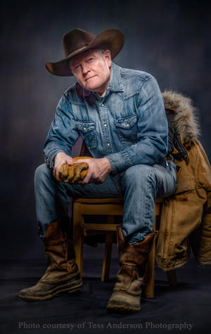 Portrait of Craig Johnson sitting a in a chare in denim jeans and shirt with muck boots